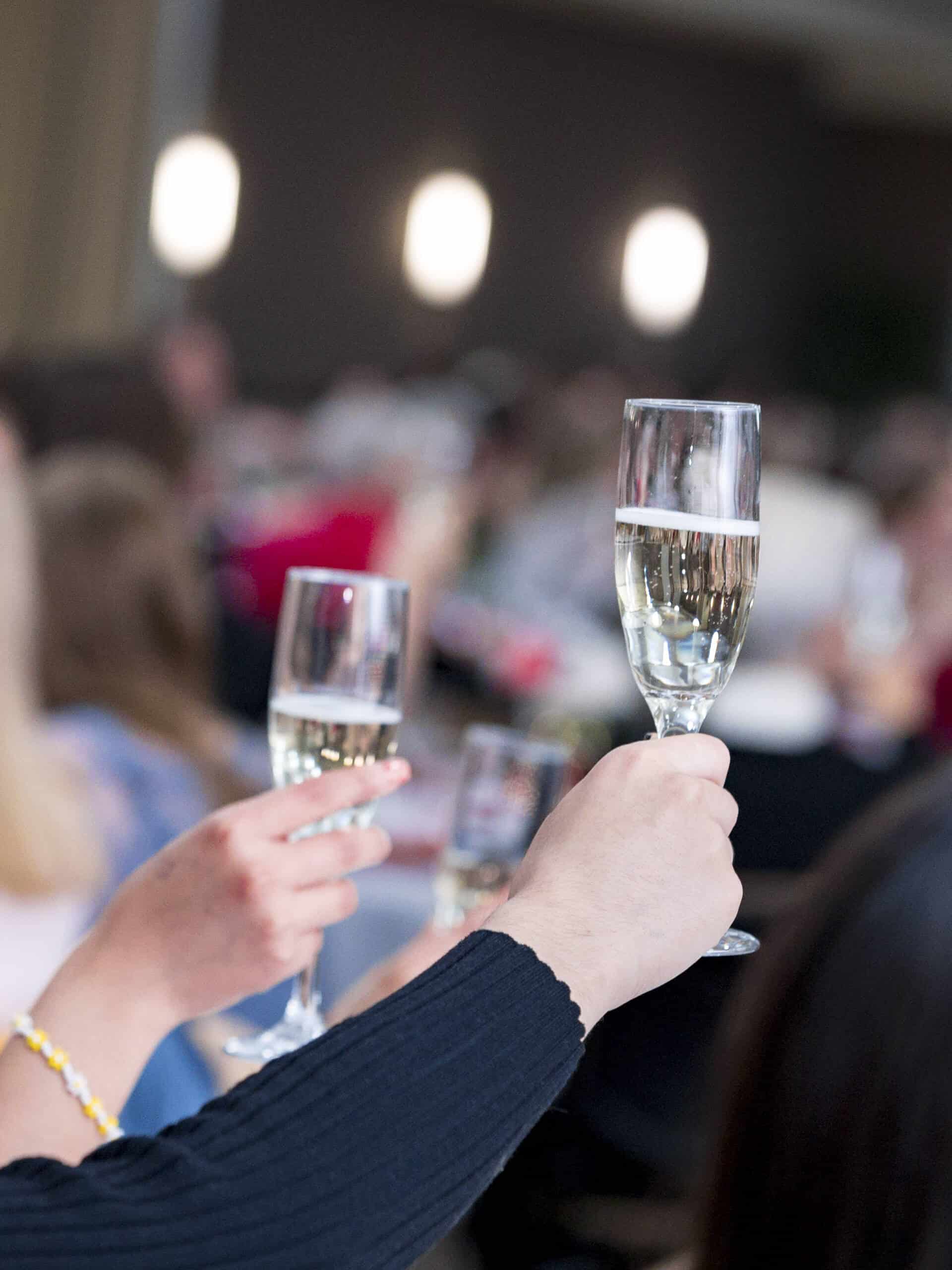 People raising glasses of champagne in a toast at a social event, with a blurred background of other attendees and soft lighting.