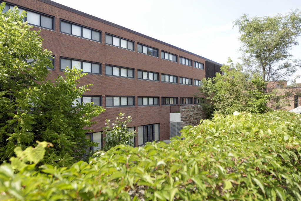 A four-story brick building with rows of windows, partially obscured by lush green trees and shrubs in the foreground, is seen on a sunny day with a clear sky.
