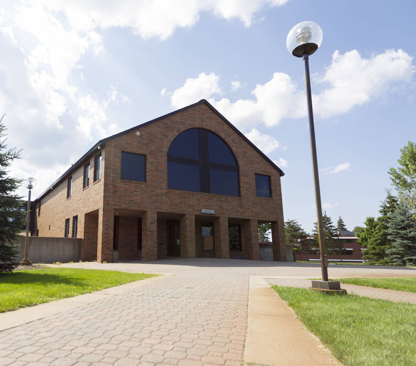 A two-story brick building with large, dark windows and an arched roof section stands under a partly cloudy sky. A pathway leads to the entrance, lined with grass, a streetlamp, and an evergreen tree to the left of the building.