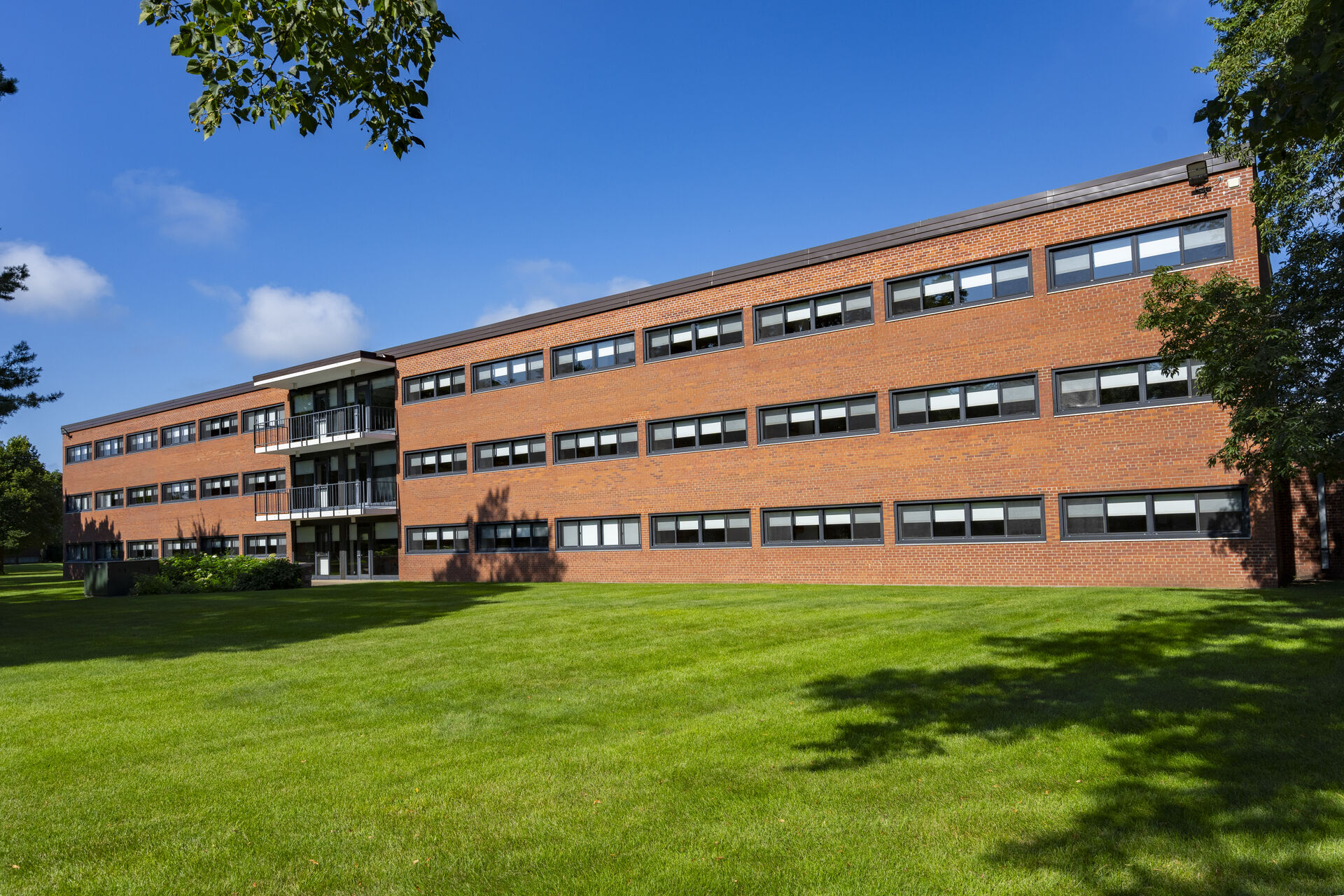 A three-story brick building with large windows, surrounded by a well-maintained green lawn and trees, under a clear blue sky.