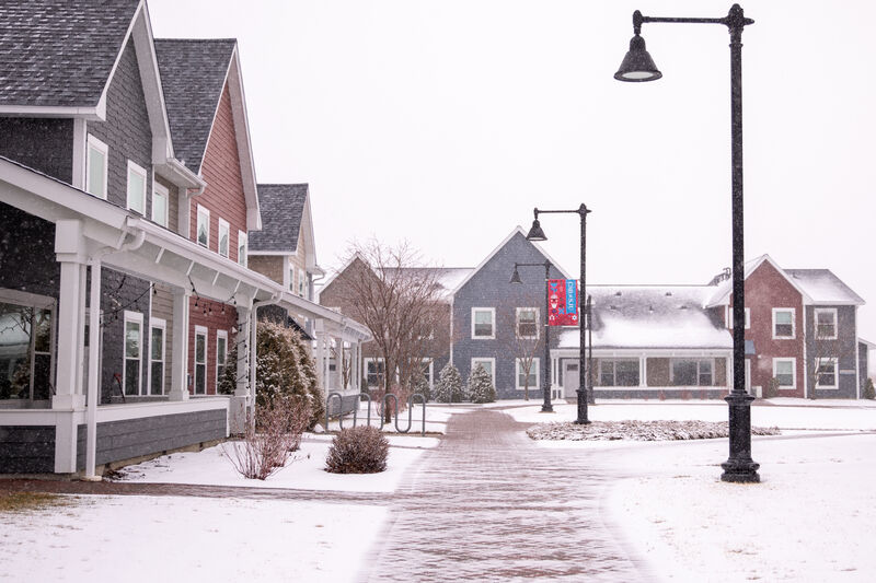 A snowy path leads between modern, colorful houses with peaked roofs on a quiet, overcast day. Light snow covers the ground and roofs, and black street lamps line the walkway.