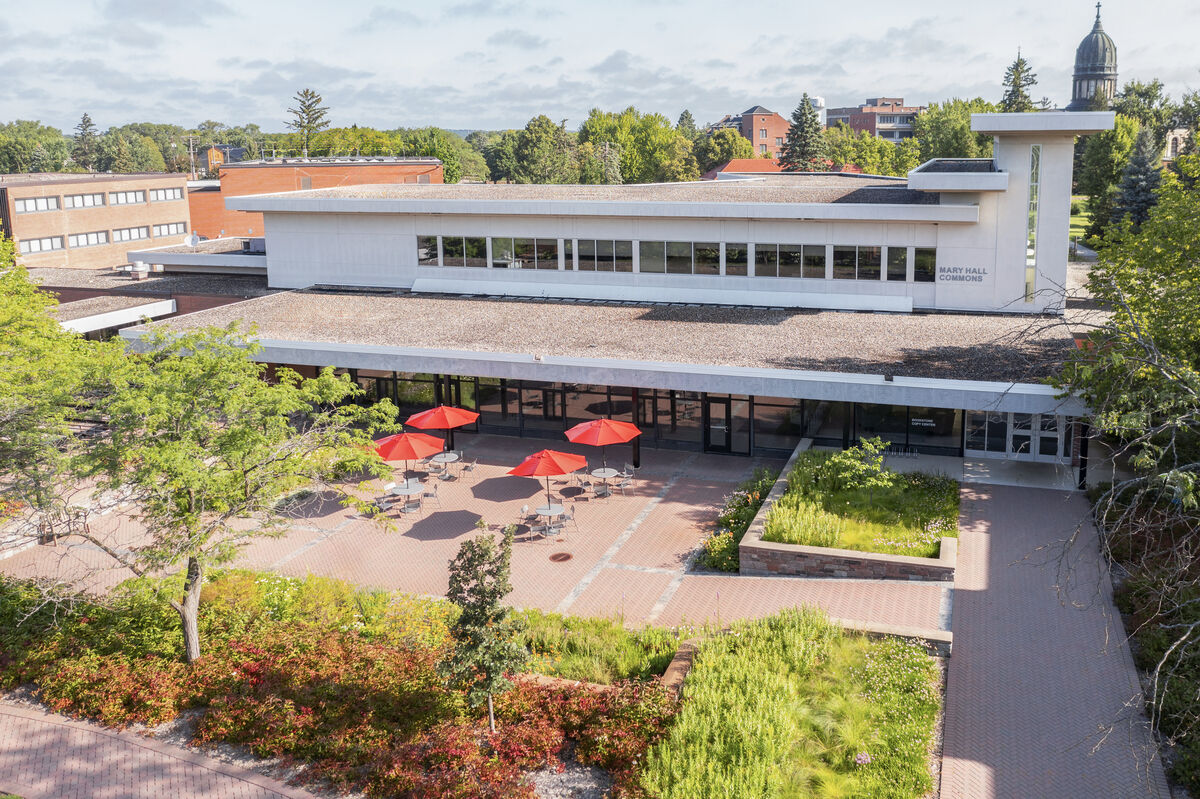 A modern white building with large windows and a flat roof sits behind a courtyard with red umbrella-covered tables, green shrubs, and trees, under a clear sky.