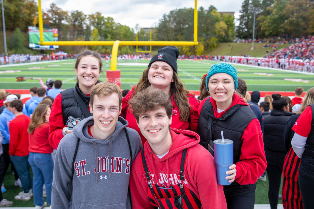 A group of five young adults, dressed in St. John's University apparel, stand smiling in front of a football field. The stadium is filled with spectators, and a football game is ongoing in the background. The weather appears cool, with some group members wearing beanies.