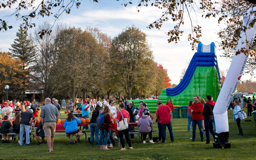 A lively outdoor gathering features people socializing and eating at picnic tables on a grassy area. A large inflatable slide is visible in the background. Trees with autumn foliage surround the scene under a clear sky.