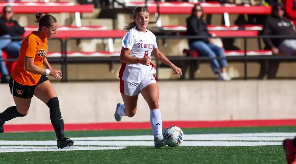 A soccer player in a white "St. Benedict" uniform dribbles the ball on a field, followed closely by an opponent in an orange jersey. Spectators sit in red bleachers in the background.