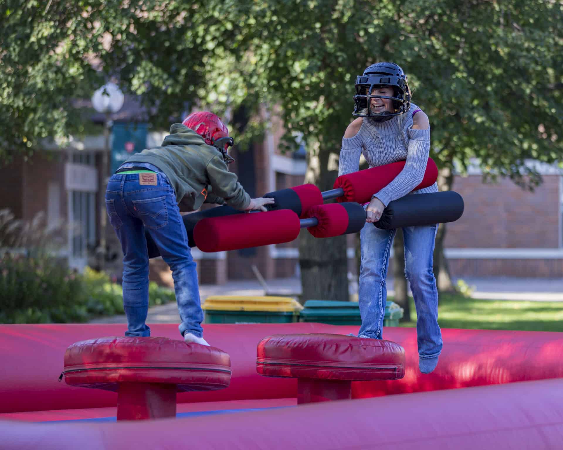 Two people wearing helmets and casual clothes balance on round red platforms in an inflatable arena, playfully jousting with large padded sticks outdoors on a sunny day.