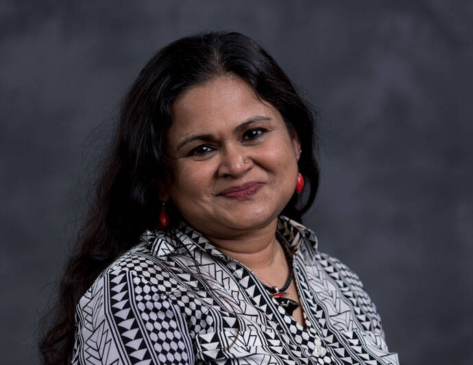 A woman with long dark hair and red earrings is smiling. She is wearing a patterned blouse with geometric designs, and the background is a gradient of dark gray shades.