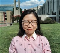 A person with medium-length dark hair and glasses smiles at the camera. They are wearing a pink shirt with a pattern. In the background, there are buildings, green grass, and a partly cloudy blue sky.