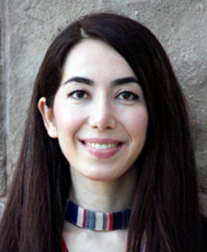 A woman with long, straight dark hair and light skin smiles at the camera. She is wearing a striped neck accessory and a red patterned top. The background is a textured brown wall.