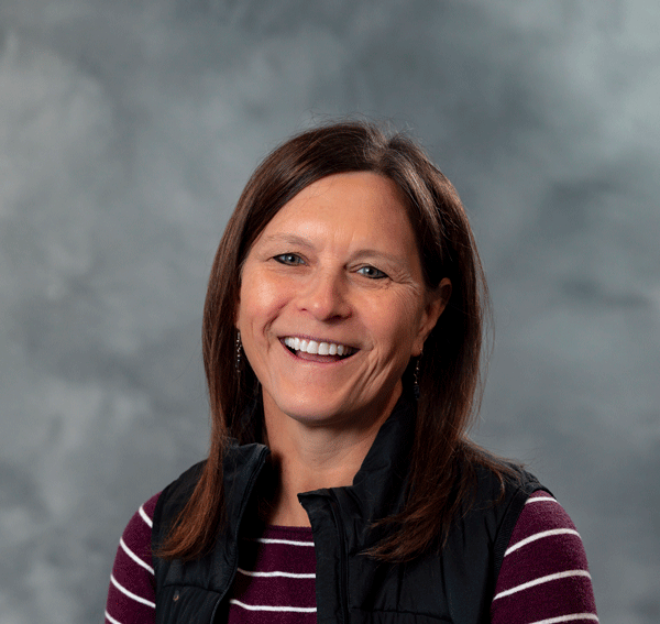 Diana Fenton, Professor. A woman with long brown hair is smiling, wearing a black vest over a maroon and white striped shirt. The background is a gray gradient.