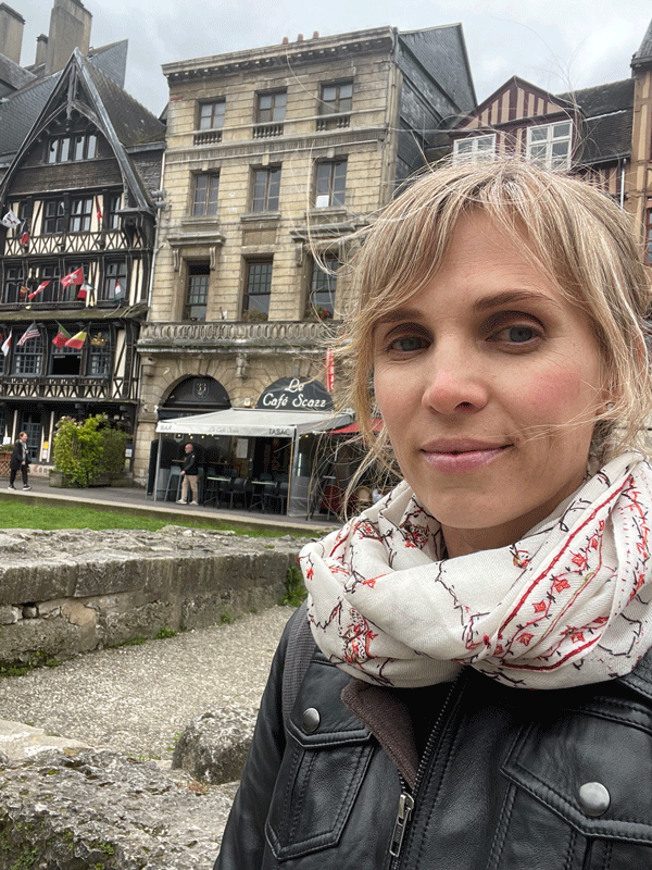 A person with a light scarf and dark jacket stands in front of historic European-style buildings. The street scene includes a café with people near the entrance. The sky is overcast.