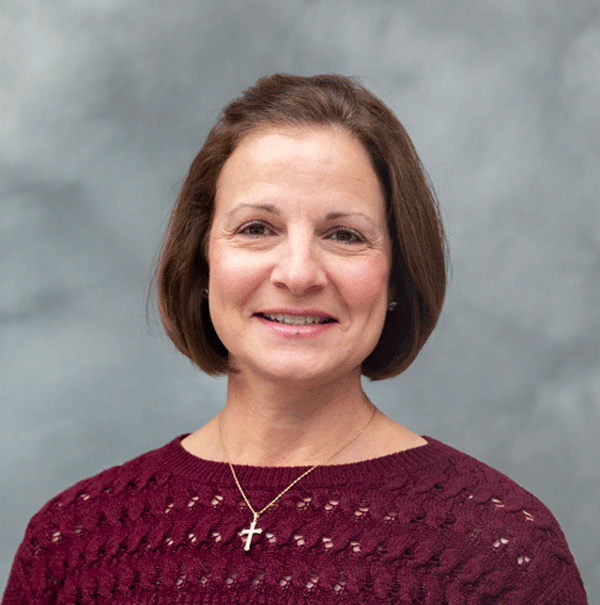 A woman with short brown hair smiles at the camera. She is wearing a maroon sweater with a cross necklace. The background is softly blurred in gray tones.
