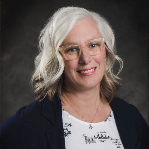 Alicia Peters, Professor. A person with shoulder-length white and gray hair, wearing glasses, a white patterned shirt, and a dark blazer smiles against a dark background.