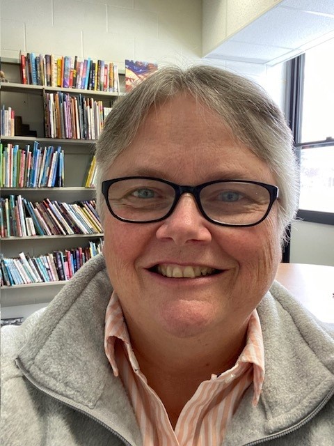 Smiling individual with short gray hair and glasses in a room with bookshelves. Wearing a gray jacket over a striped shirt, standing in front of a window with natural light.