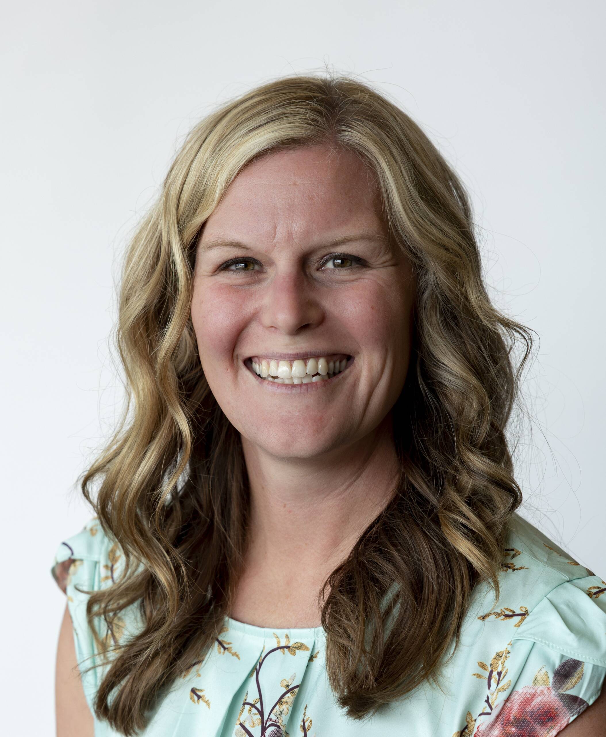 A woman with wavy blonde hair and a floral light blue top smiles against a plain white background.