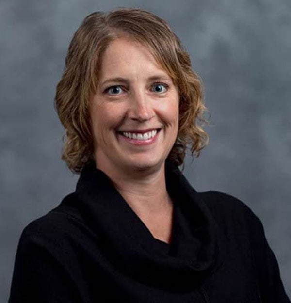 A woman with short, wavy, light brown hair is smiling at the camera. She is wearing a black top and is posed against a gray, blurred background.