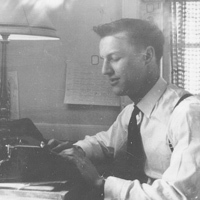A black and white photo of a man sitting at a desk, typing on a typewriter. He is wearing a dress shirt, tie, and suspenders. There is a lamp on the desk and papers pinned on the wall in the background. Light filters through a window with blinds to the right.