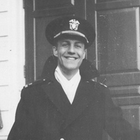 A smiling person in a naval uniform and hat stands in front of a building. The image is in black and white, and the individual appears to be posing for the photograph.