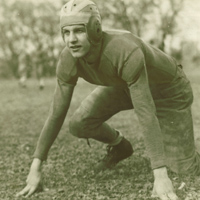 A vintage black-and-white photograph of an American football player in an old-fashioned helmet and padded uniform, crouching in a ready stance on a grassy field. Trees are visible in the blurred background.