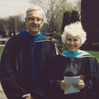 An elderly man and woman, both wearing academic gowns and mortarboards, stand outdoors on a sunny day. They are smiling and holding folders, with trees and a brick building visible in the background.