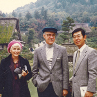 Three people standing outdoors with a serene, wooded landscape and traditional buildings in the background. One woman is wearing a pink beret, and two men are dressed in gray jackets. One of the men is wearing a blue cap and eyeglasses.