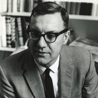 A grayscale photo shows a man wearing dark-rimmed glasses, a suit jacket, a collared shirt, and a tie. He is seated and appears to be in a library or office, with shelves of books in the background. The man is looking off to his left.