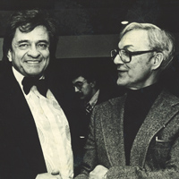 Two men engaged in conversation at an event. One man, on the left, wears a bow tie and the other man, on the right, wears glasses and a suit jacket. Both are smiling. A third person is visible in the background. The image is in black and white.