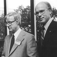 Black and white photo of two men in suits and glasses. One man looks down while holding papers, and the other faces the camera with a slight smile. Trees and a window are visible in the background.