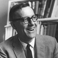 A black-and-white photo of a smiling person wearing glasses, a suit, and a tie. They are in an indoor setting with a bookshelf filled with books in the background.