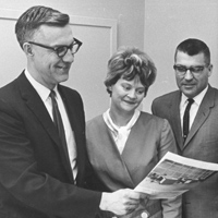 Three people, two men and one woman, are standing together and smiling while looking at a document held by the man on the left. All are dressed in formal attire, and they appear to be in an office setting.