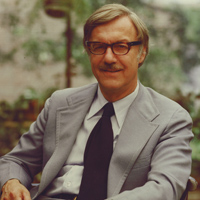 A man with gray hair and glasses is sitting outdoors. He is wearing a light gray suit, a white dress shirt, and a black tie. He is smiling gently, and the background features blurred greenery.