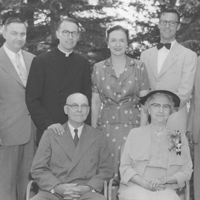A black-and-white photo of seven people. Two elderly people sit in front, one man and one woman. Five adults stand behind them, including three men (one in clerical attire) and two women. They all pose outdoors in front of trees, smiling at the camera.