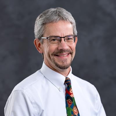 A smiling man with short gray hair and glasses is pictured against a dark background. He is wearing a white dress shirt and a colorful tie featuring cartoon faces.