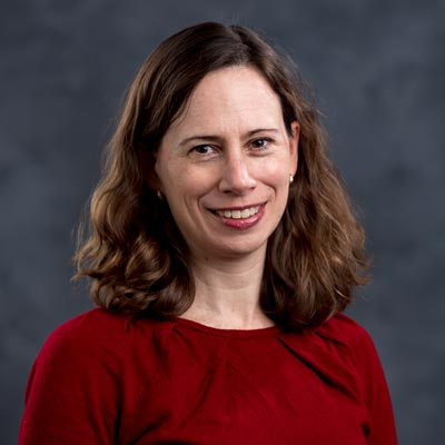 A woman with shoulder-length brown hair is smiling at the camera. She is wearing a red top and small earrings. The background is a dark, neutral color.