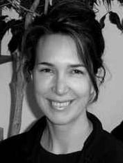 Black and white photo of a smiling person with their hair pulled back, standing in front of a plant. They are wearing a dark top.