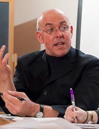 A man with glasses, wearing a black suit, is speaking while gesturing with his hands. He holds a pen in one hand, and another person next to him holds a purple pen, visible in the foreground. They appear to be engaged in a discussion.