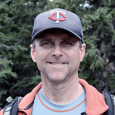 A man wearing a baseball cap with a red "C" and "T" logo, a light blue shirt, and a red jacket stands outdoors, smiling at the camera. The background consists of green foliage and trees.