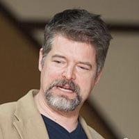 An individual with short brown hair, a beard, and a mustache is looking slightly to the side. They are wearing a beige blazer over a dark shirt. The background is blurred and minimal.