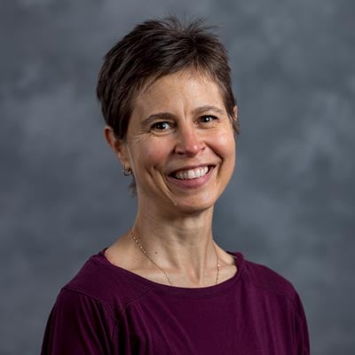 A person with short dark hair smiles while looking at the camera. They are wearing a purple top and standing against a mottled gray background.