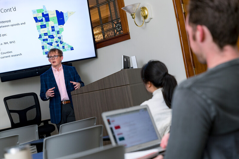 A person stands at the front of a classroom giving a presentation. A large screen behind them displays a map. Two people in the foreground look toward the presenter, one using a laptop. The room has wooden accents and a sconce on the wall.