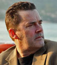 A man with short dark hair and a trimmed beard is looking off to the side. He is wearing a beige blazer and appears contemplative. The background is slightly blurred, showing a body of water and distant landscape.