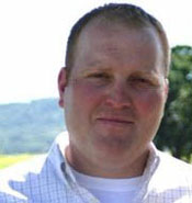 A person with short, light hair stands outside. They are wearing a light-colored, button-up shirt. The background is an outdoor setting with trees and a distant hill visible under a clear sky.