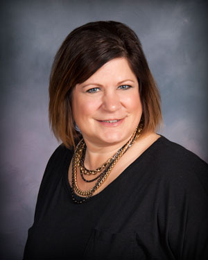A woman with shoulder-length brown hair smiles at the camera. She is wearing a black top and a chunky gold necklace. The background is a muted blend of dark and light tones, giving a professional studio portrait feel.