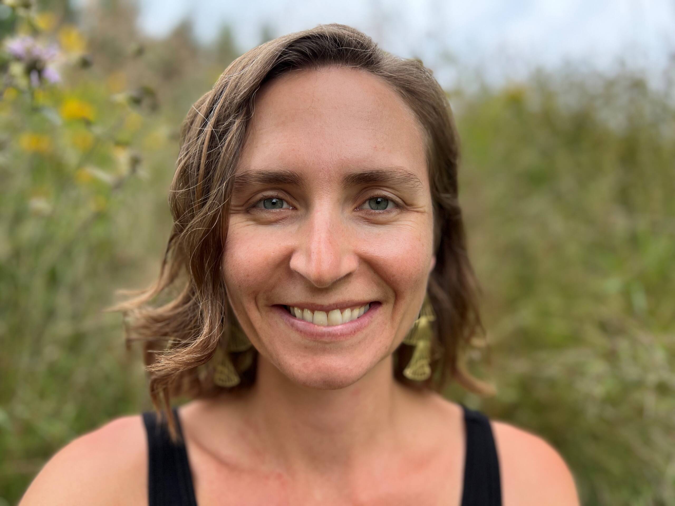 A person with shoulder-length wavy hair smiles at the camera, standing outdoors with a blurred background of green foliage and wildflowers. They are wearing a black sleeveless top and large earrings.