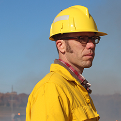 A man wearing glasses and a yellow hard hat stands outdoors against a blue sky. He is dressed in a yellow safety jacket and looks focused, with a hazy background indicating possible industrial or construction activity.