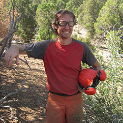 A person wearing glasses and an orange and gray shirt stands outdoors in a wooded area, smiling. They are holding an orange helmet and have their other arm resting on a tree branch. The background features green trees under bright sunlight.