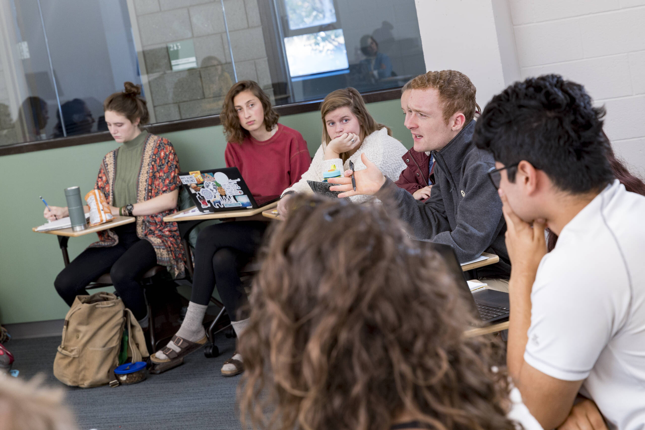 A group of students sits in a classroom, engaged and attentive. One student in the middle speaks passionately, gesturing with his hands, while others listen thoughtfully. Some have laptops or notebooks in front of them. The room has desks arranged in a semi-circle.