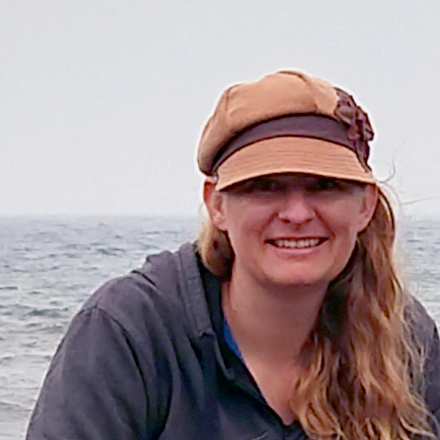 A woman with long, wavy hair wearing a beige newsboy cap with a flower detail and a dark hoodie smiles while standing near the ocean on a cloudy day.