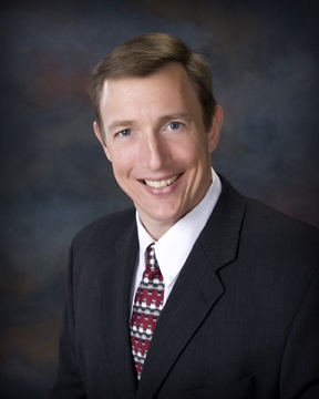 A man with short brown hair, wearing a black suit, white shirt, and patterned red and white tie, smiles at the camera. The background is a blurred, dark gradient.