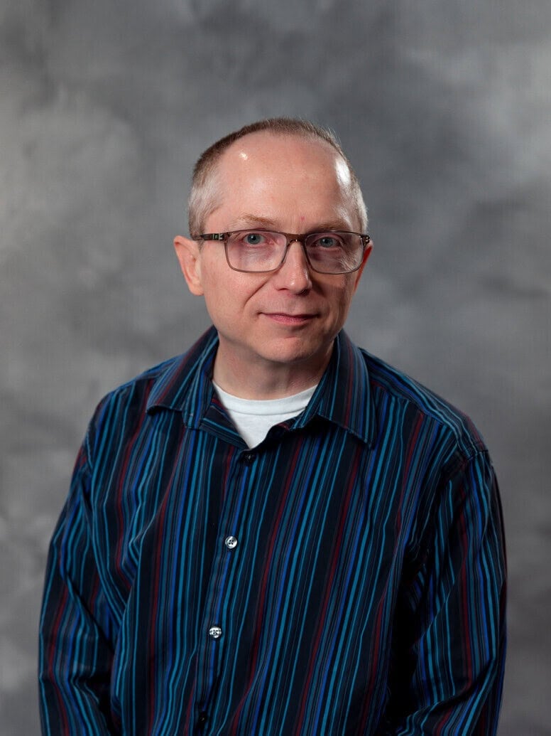 A middle-aged man with short hair and glasses wears a blue, black, and red striped shirt and white t-shirt, posing in front of a gray, cloudy backdrop. He has a neutral expression and is looking at the camera.